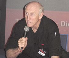 waist-high portrait wearing black shirt, holding microphone, leaning forward and speaking